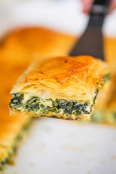 A square piece of freshly baked spanakopita being held up on a large silver spatula.