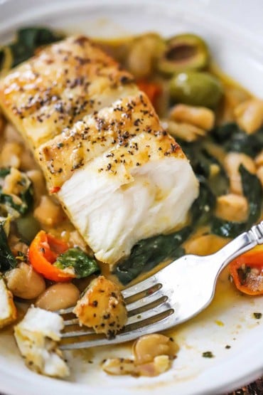 A close-up view of a seared fillet of halibut that has a bite missing and is sitting on a bed of sautéd spinach, tomatoes, white beans, and broth.