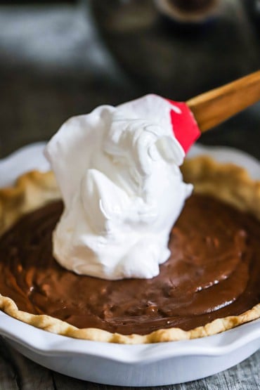 A close-up view of a mound of white meringue being transferred from a red spatula on of a chocolate pie.