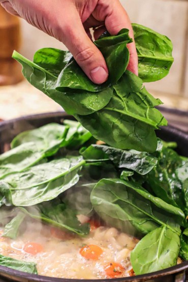 A person dropping fresh spinach leaves into a skillet of a simmering bean and tomato sauce.