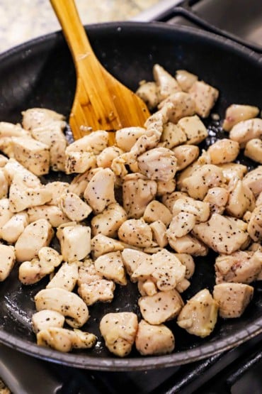 A large black nonstick skillet filled with bite-sized pieces of chicken that have been fully cooked with a spatula sitting off to the side.