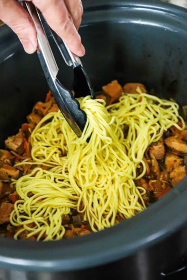A person using a pair of tongs to place cooked chow mein noodles into a slow-cooker filled with a chicken and vegetable mixture.