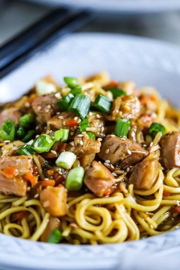 A close-up view of a bowl filled with slow-cooker chicken chow mein topped with sliced scallions and toasted sesame seeds.