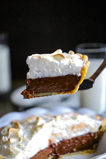 A slice of chocolate meringue pie being raised up by a metal pie slicer over the pie itself.