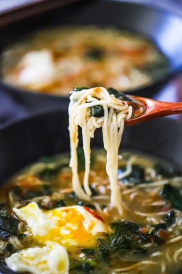 A small wooden spoon being used to lift ramen noodles out of a bowl filled with ramen soup with spinach and a broken open poached egg.
