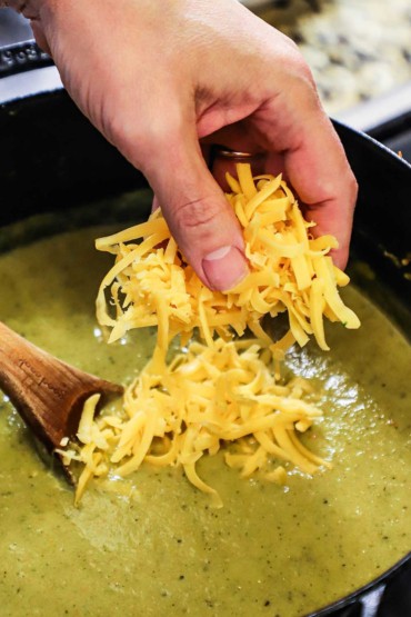 A person dropping a large handful of shredded cheddar cheese into a pot of puréed broccoli soup.