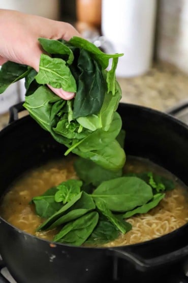A person dropping fresh spinach into a black pot filled with chicken broth and ramen noodles.