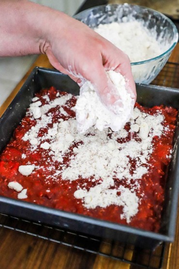 A person sprinkling a flour and cubed butter mixture over a metal pan filled with a raspberry filling on top of a baked shortbread crust.