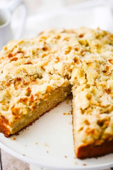 An apple cake on a white circular cake dish with a triangular slice of the cake missing