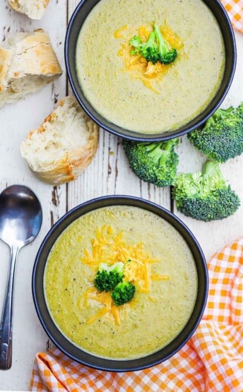An overhead view of two black soup bowls filled with broccoli cheddar soup next to pieces of bread and uncooked broccoli florets.