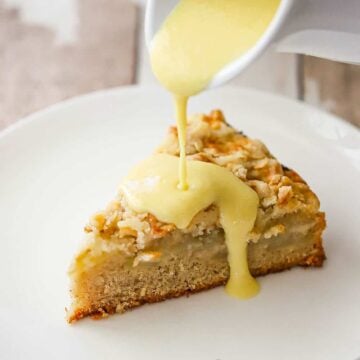 Vanilla custard being poured from a small white pitcher over a slice of Irish apple cake on a white dessert plate.