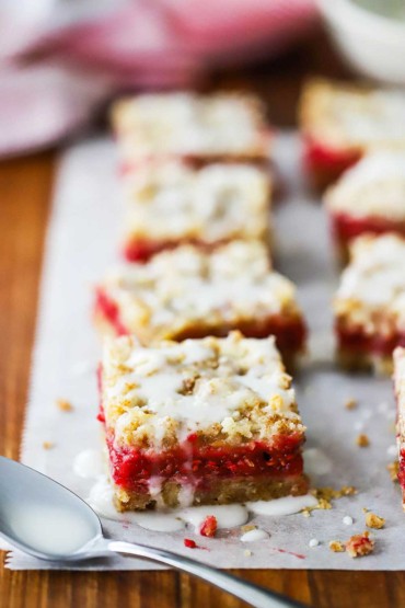 Two rows of square raspberry Linzer bars sitting on parchment paper with a vanilla glazed drizzled over the top.