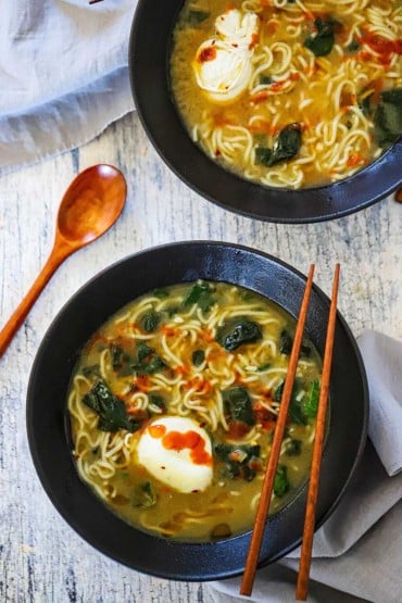 An overhead view of two ramen soup bowls filled with ramen with spinach and poached eggs topped with sriracha sauce.