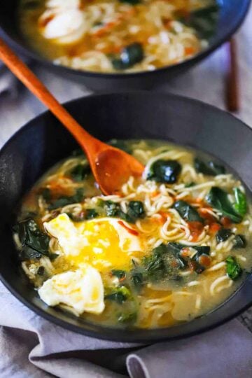 A close-up view of a black soup bowl filled with ramen with spinach and poached egg with a wooden spoon inserted into it.