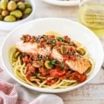 A shallow pasta bowl filled with a serving of salmon puttanesca sitting on a bed of bucatini.