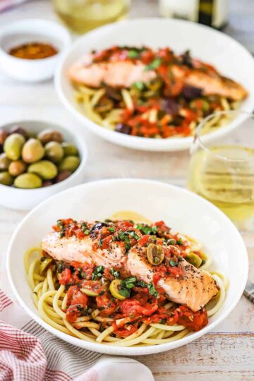 An overhead view of a two shallow pasta bowls each filled with a bed of bucatini pasta topped with salmon and a puttanesca sauce.