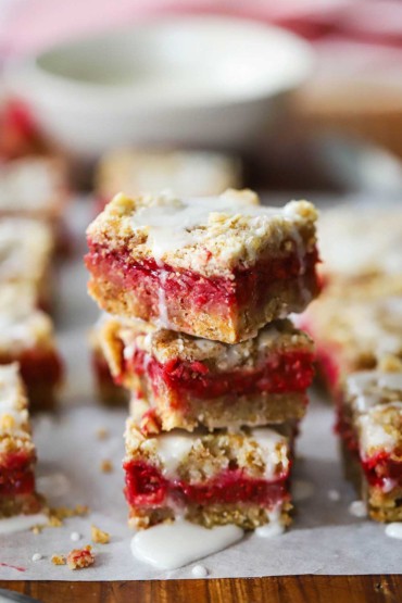 A close-up view of three raspberry Linzer bars stacked on top of each other on a sheet of parchment paper with other Linzer bars nearby.