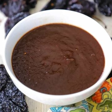 A white bowl filled a brown ancho chili sauce that is sitting next to dried ancho chilils and a festive colorful napkin.