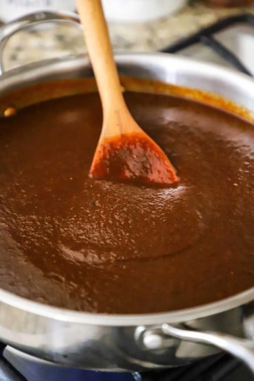 A wooden spoon inserted into the middle of a stainless steel skillet filled with a smooth brown enchilada sauce.