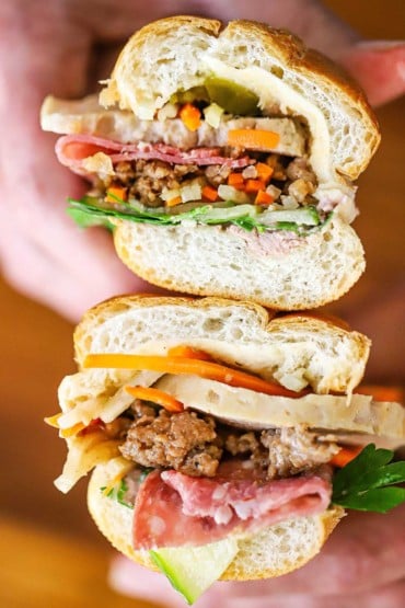 A person holding a banh mi sandwich that has been sliced in half so the components of the sandwich are visible including pork, vegetables, and herbs.