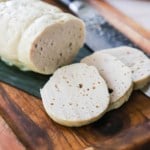 Three slices of cha lua sitting on a cutting board lined with a banana leaf and a loaf of the cha lua nearby.
