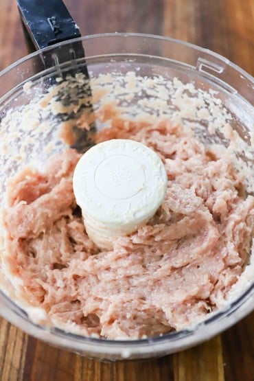 An overhead view of ground pork in a large food processor that has been combined with seasonings and water.