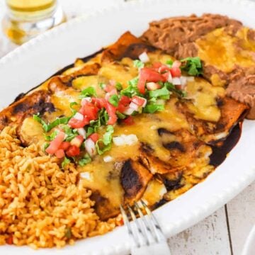A close-up view of a large white platter filled with Mexican rice on one side and refried beans on the other side and three cheese enchiladas with ancho sauce in the middle.