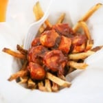 A straight-on view of a basket lined with white parchment paper that is filled with a helping of German currywurst with a plastic white fork stuck in the side.