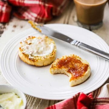 Two homemade English muffin halves covered with butter and peach jam with one that has a bite taken out of it.