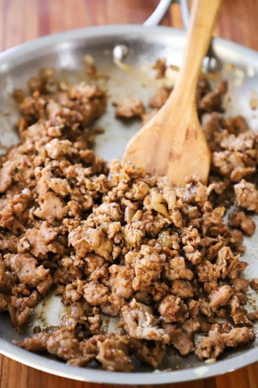 A close-up view of ground pork that has been cooked and seasoned with Asian spices in a steel skillet with a wooden spatula in it.