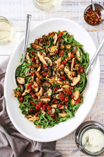 An overhead view of a large white platter filled with spinach pasta and wild mushroom sauce with two serving spoons inserted on the sides.