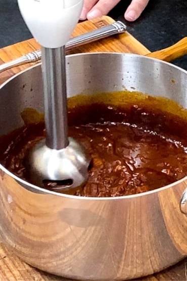 A person using an immersion blender to purée a curry tomato sauce in a silver saucepan.