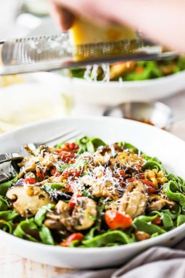A person grating a block of Parmesan cheese over a pasta bowl filled with spinach linguine with sautéed wild mushrooms and tomatoes.