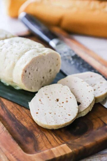 Three slices of cha lua sitting on a cutting board lined with a banana leaf and a loaf of the cha lua nearby.