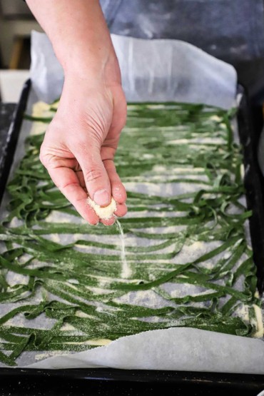 A person sprinkling semolina flour over strips of fresh spinach linguine on parchment paper on a baking pan.