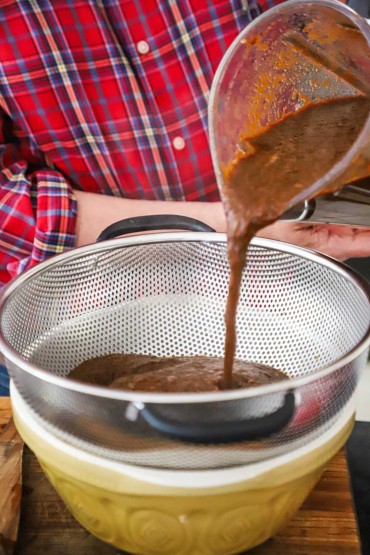 A person pouring a brown chili sauce from a blender into a fine-mesh colander over a heat-proof bowl.