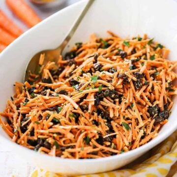 An oval white serving bowl filled with carrot and raisin salad with a gold serving spoon inserted on the side of the bowl.