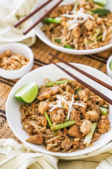 A straight-on view of a white bowl filled with chicken pad Thai topped with bean sprouts, scallions, and a lime wedge off to the side.