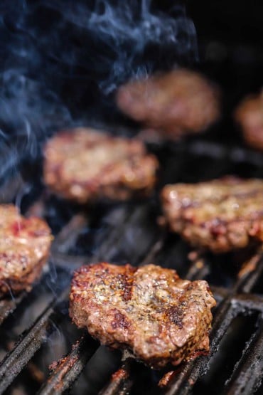 Several beef slider patties on a hot gas grill with smoke coming from below the grates.