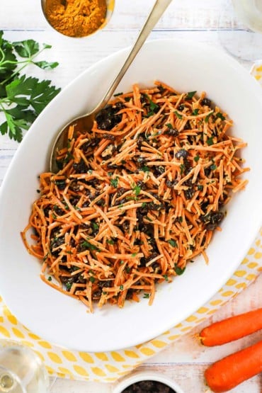 An overhead view of an oval white serving bowl filled with carrot and raisin salad with curry dressing.