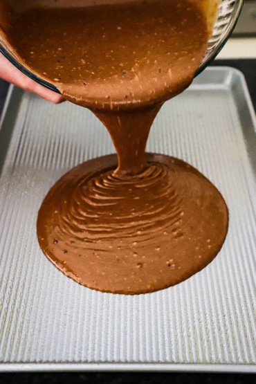 A liquid chocolate cake batter poured from a glass bowl into a half sheet pan.