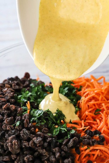 A creamy curry dressing being poured over a bowl filled with raisins, chopped parsley, and strips of fresh carrots.