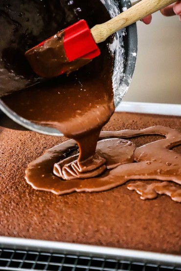 Warm chocolate frosting being transferred with a red spatula from a glass bowl onto the top of a chocolate Texas sheet cake in a sheet pan on a baking rack.