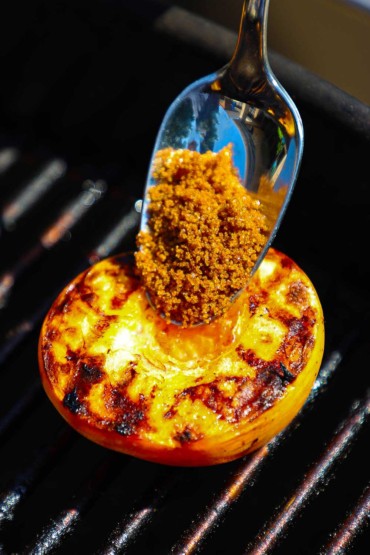 A person using a silver spoon to place brown sugar on top of a halved peach that is on a hot charcoal grill.
