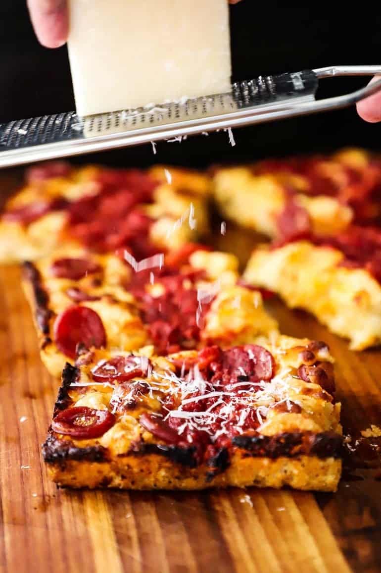 A straight-on view of a person grating a block of Romano cheese over a slice of Detroit-style pizza with pepperoni on top.