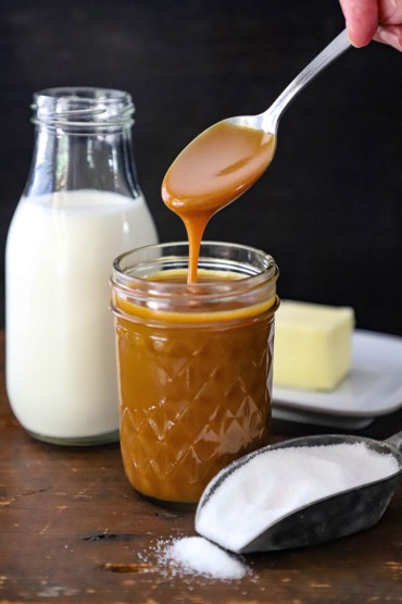 A person pulling a small spoon out of a jar of caramel with caramel dripping from the spoon back into the jar.