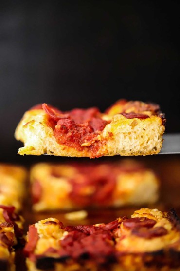 A slice of Detroit-style pizza being raised above the pizza with a metal spatula.