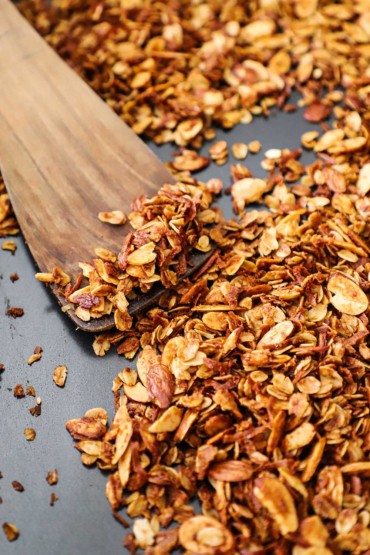 A close-up view of a pan that is filled with homemade granola and a wooden spatula inserted into the middle of it.