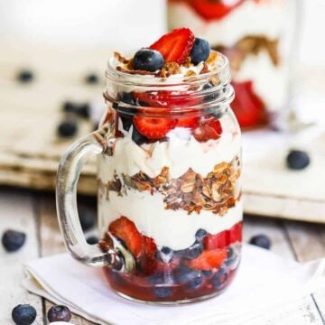 A straight-on view of a glass jar with a handle holding a wild berry parfait with mascarpone and topped with strawberries, blueberries, and granola.