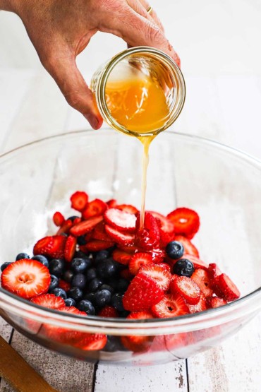 A person pouring an orange-honey dressing from a small jar into a large bowl filled with fresh cut fruit.